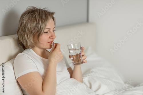 a middle-aged white woman with moisturized beautiful skin holds a glass of clean water and vitamins in her hands while sitting on the bed. the concept of health and beauty care