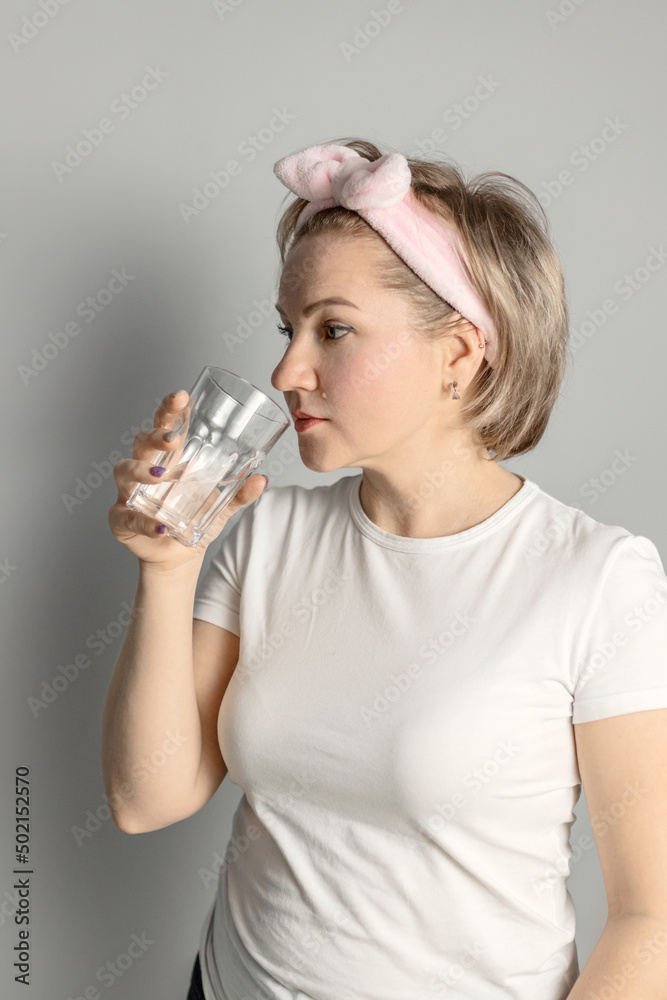 a middle-aged woman in a white T-shirt with a headband drinks water from a glass against a gray wall a middle-aged woman in a white T-shirt with a headband drinks water from a glass against a gray wall