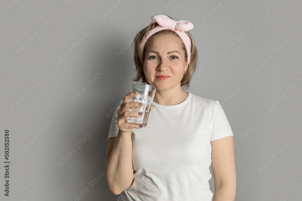 a middle-aged woman in a white T-shirt with a headband drinks water from a glass against a gray wall a middle-aged woman in a white T-shirt with a headband drinks water from a glass against a gray wall