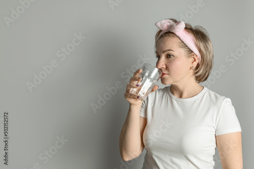 a middle-aged woman in a white T-shirt with a headband drinks water from a glass against a gray wall