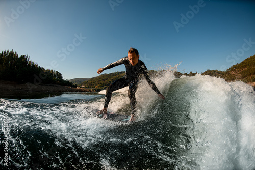 athletic man in wetsuit on wakesurf board riding down the splashing wave