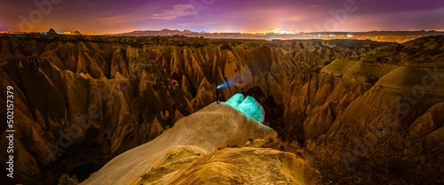 Panoramic view of red valley in Cappadocia at night with stars in the background and person in the middle surounded by rock formations. Astrophotography in Turkey. Background blank space image.