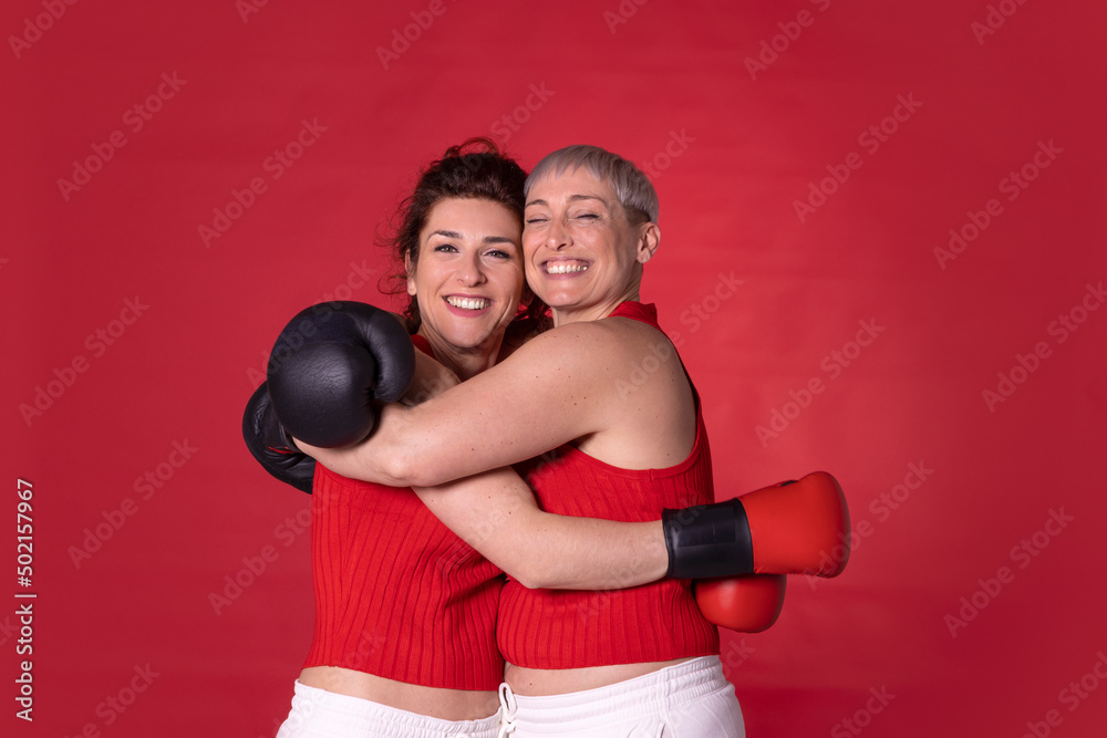 Smiling friends wearing boxing gloves hugging each other against red background Smiling friends wearing boxing gloves hugging each other against red background
