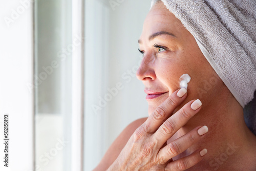 Smiling woman applying moisturizer on face at home