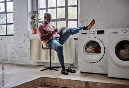Mature man using tablet PC with foot on washing machine sitting on chair at home