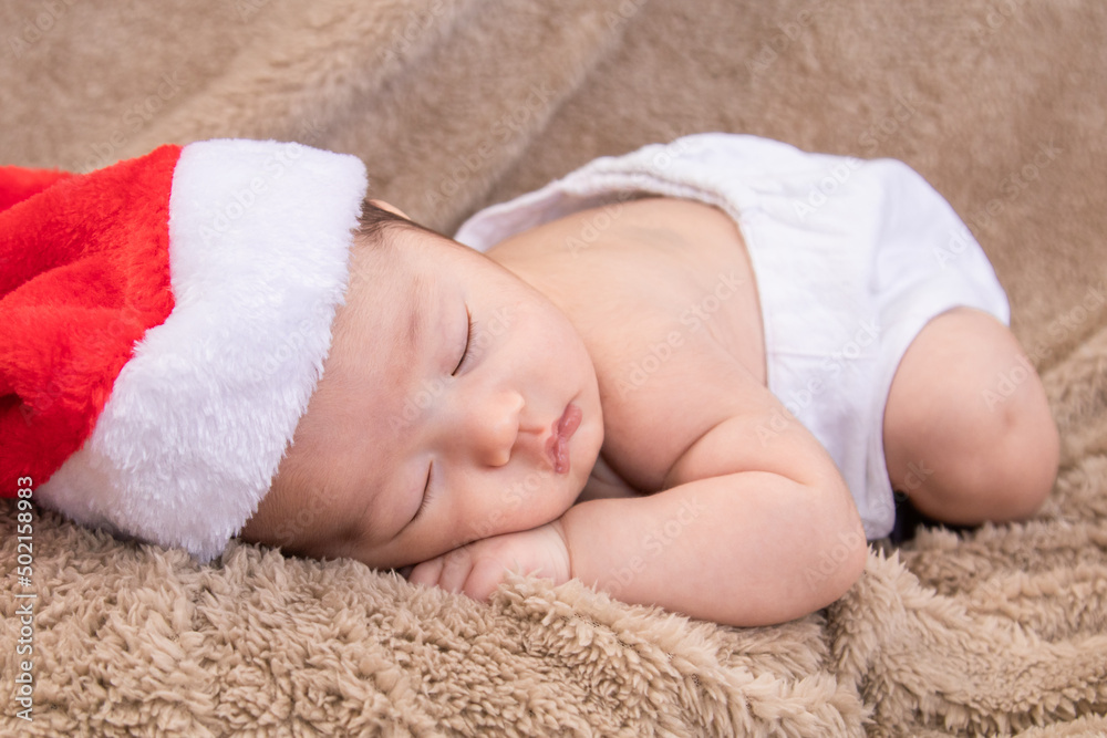 Asian newborn baby wearing christmas hat sleeping on brown blanket, adorable infant lying on bed at home with trust and safe. Little child was born on the same day as Jesus. Asian newborn baby wearing christmas hat sleeping on brown blanket, adorable infant lying on bed at home with trust and safe. Little child was born on the same day as Jesus.