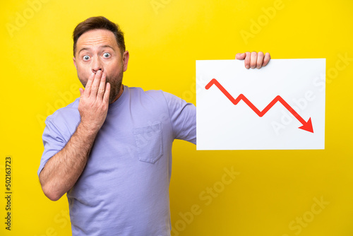 Middle age caucasian man isolated on yellow background holding a sign with a decreasing statistics arrow symbol with surprised expression