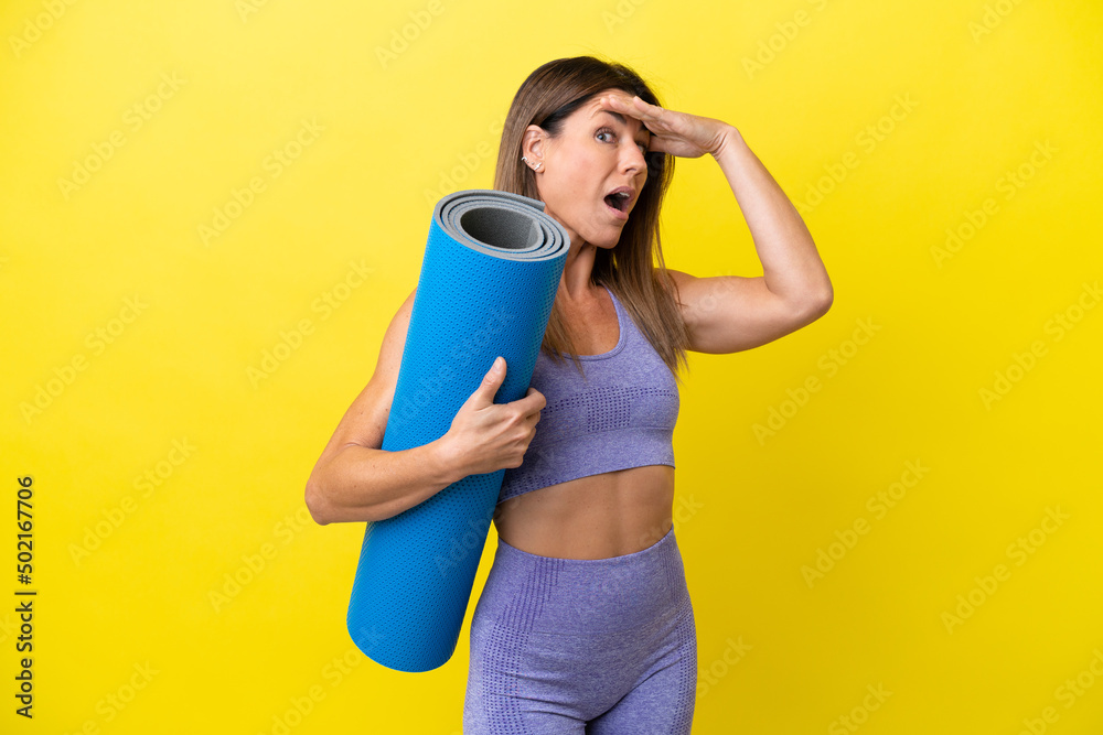 Sport woman going to yoga classes while holding a mat isolated non yellow background doing surprise gesture while looking to the side Sport woman going to yoga classes while holding a mat isolated non yellow background doing surprise gesture while looking to the side