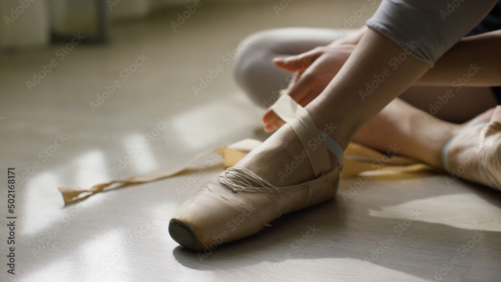 Close-up of ballerina's feet. Ballerina preparing for training, and tying ribbon of pointe shoes sitting on floor in bright sunlight