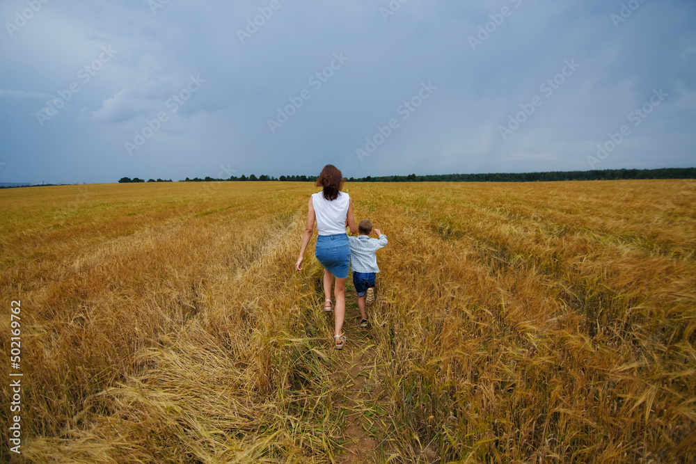 A happy family of mother and son in a summer wheat field against a stormy sky. They run across the field with their backs to the camera. Rural landscape.