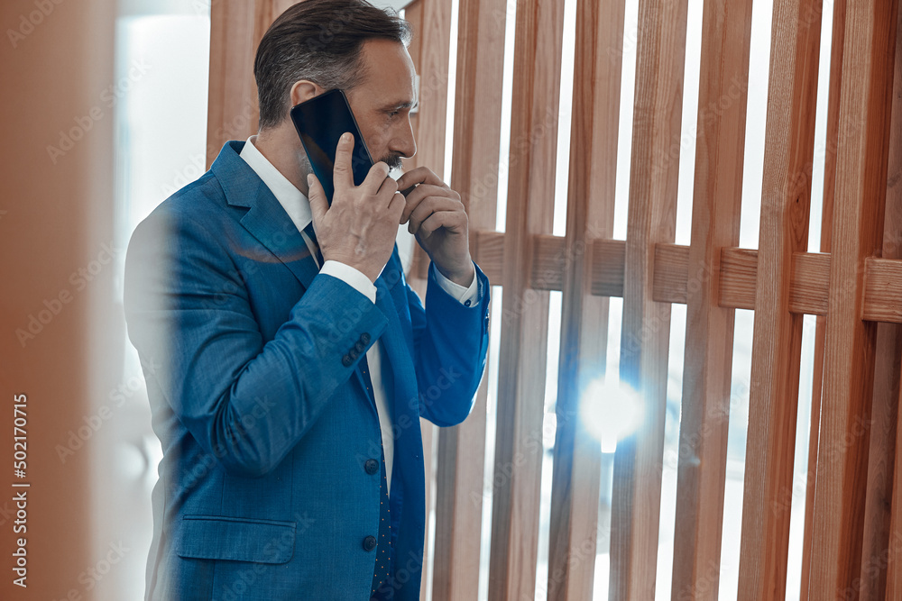 Side view of busy man in suit standing in the office space and talking on smartphone Side view of busy man in suit standing in the office space and talking on smartphone
