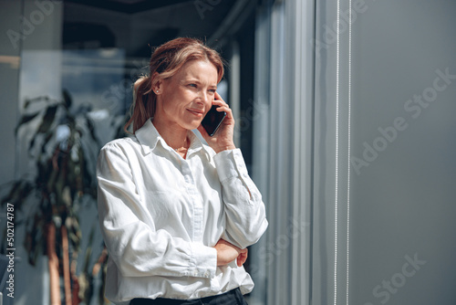 Mature female pensive businesswoman is talking on a phone and looking aside near big glass in office