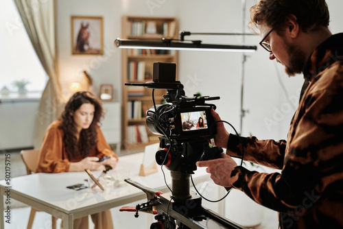 Young bearded cameraman standing in front of camera and adjusting it while shooting female blogger or advert model for commercial