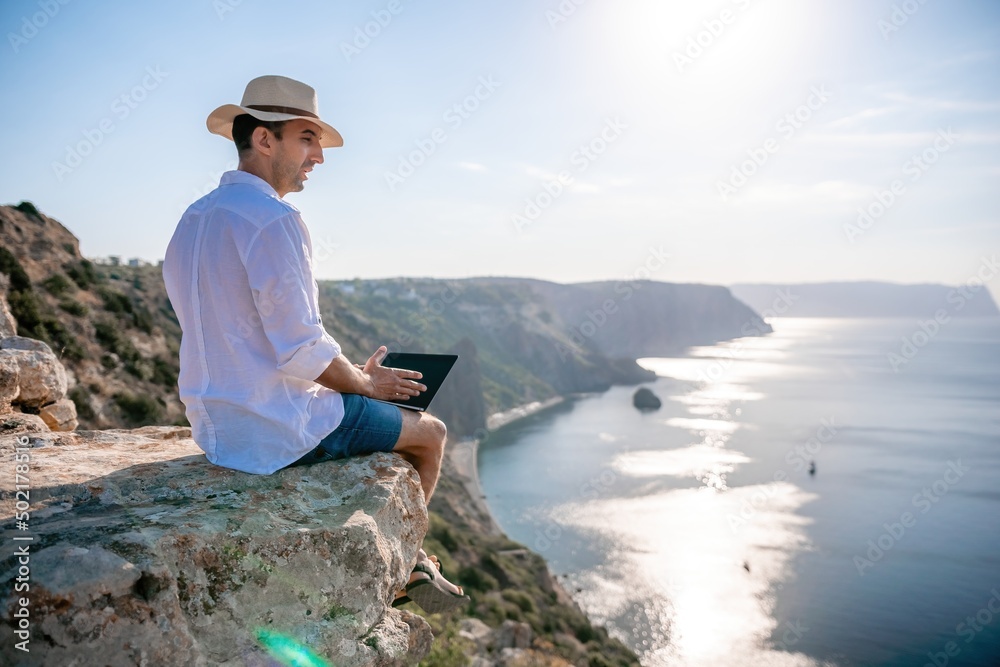 Digital nomad, man in the hat, a businessman with a laptop sits on the rocks by the sea during sunset, makes a business transaction online from a distance. Remote work on vacation.
