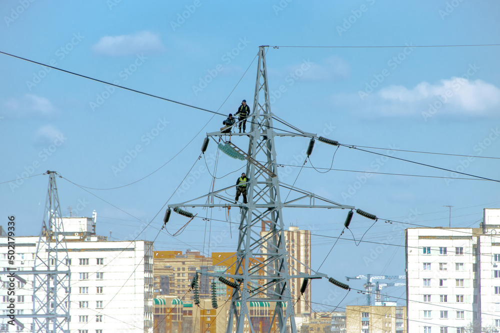High altitude installers work at on a high-voltage line pylon against the backdrop of residential buildings in a big city