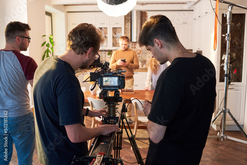 Rear view of videographer and his assistant adjusting video camera for shooting of commercial or cooking masterclass in the kitchen photo