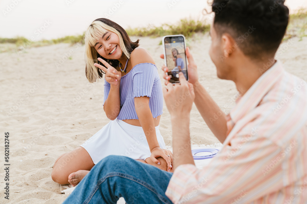 Young man taking photo of his girlfriend during picnic on beach