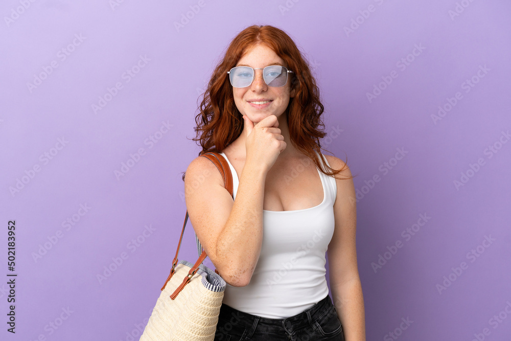 Teenager redhead girl holding a beach bag isolated on purple background with glasses and smiling Teenager redhead girl holding a beach bag isolated on purple background with glasses and smiling