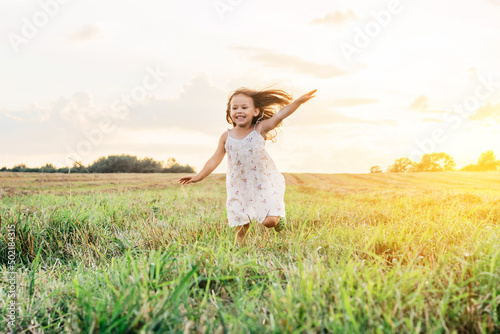 Portrait of smiling girl playing, jumping and running on grass hay field paths of dry grass in the sunset. Waving hands. Forest on bright light background. Cloudy sunny sky. Haying time