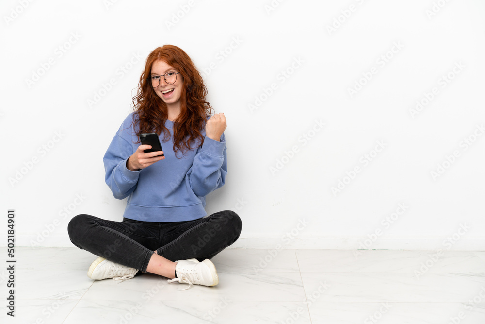 Teenager redhead girl sitting on the floor isolated on white background with phone in victory position Teenager redhead girl sitting on the floor isolated on white background with phone in victory position