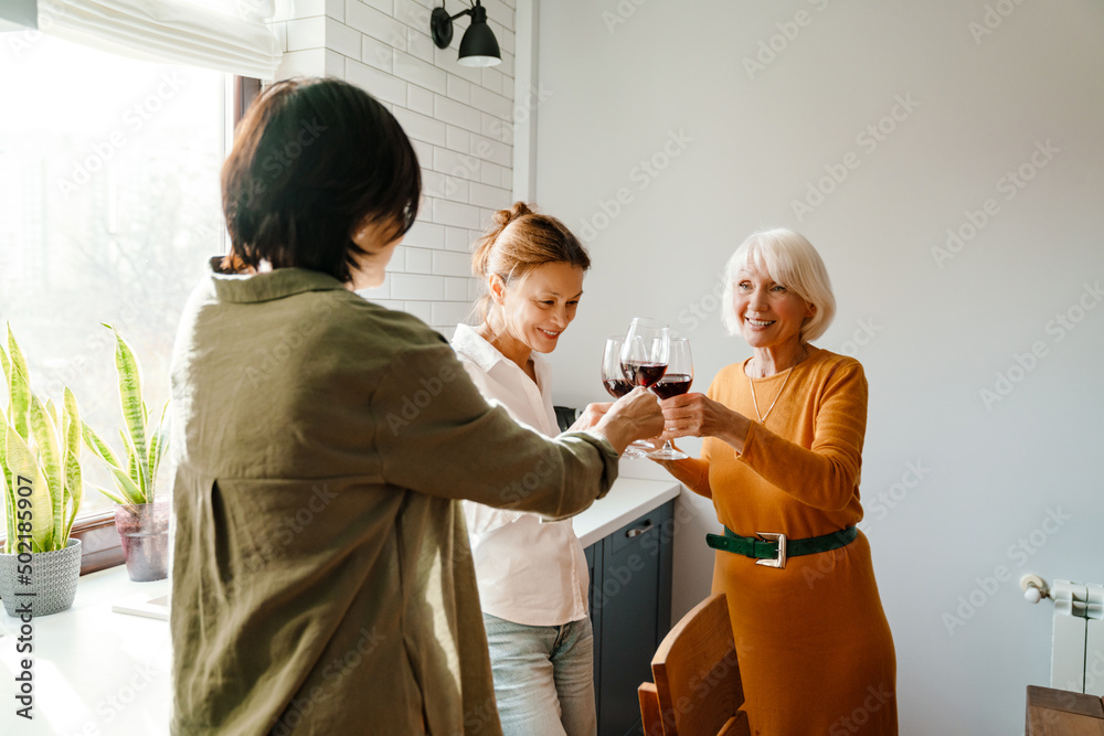 Naklejka premium Mature three women talking and laughing while drinking wine in kitchen