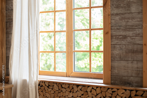 Big wooden window with frame and window sill and nature on background. Empty room, wooden window with with White linen curtain and logs decoration wall on a sunny day indoor shot. Scandinavian room