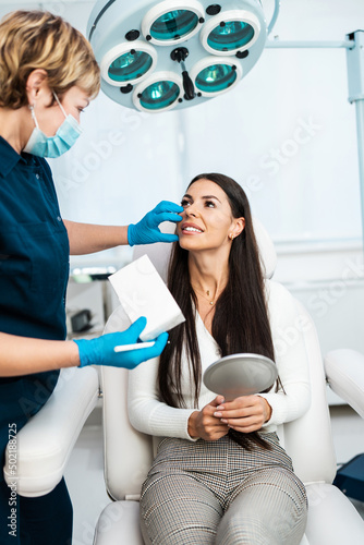 Beautiful and happy brunette woman at beauty medical clinic. She is sitting and talking with female doctor about face aesthetic treatment.