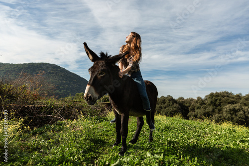 A young woman sitting on a donkey looking up at the sun in the meadow.