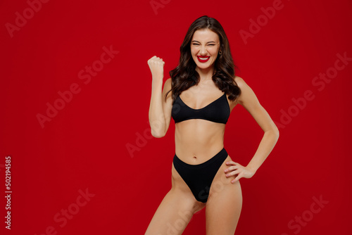 Young sexy overjoyed happy brunette woman 20s with perfect fit body wear black underwear do winner gesture clench fist isolated on plain red background studio portrait. People female beauty concept