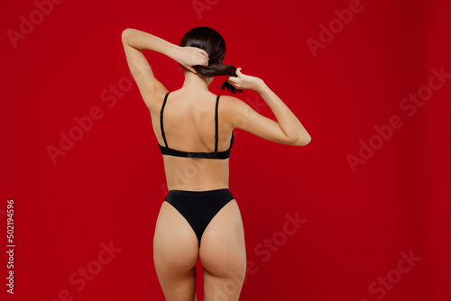 Back rear view young sexy brunette caucasian woman 20s with perfect fit body wear black underwear hold hair in ponytail isolated on plain red background studio portrait. People female beauty concept photo