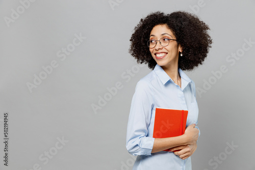 Young fun employee business corporate lawyer woman of African American ethnicity in classic formal shirt work in office hold in hand book, notebook look aside on workspace isolated on grey background.