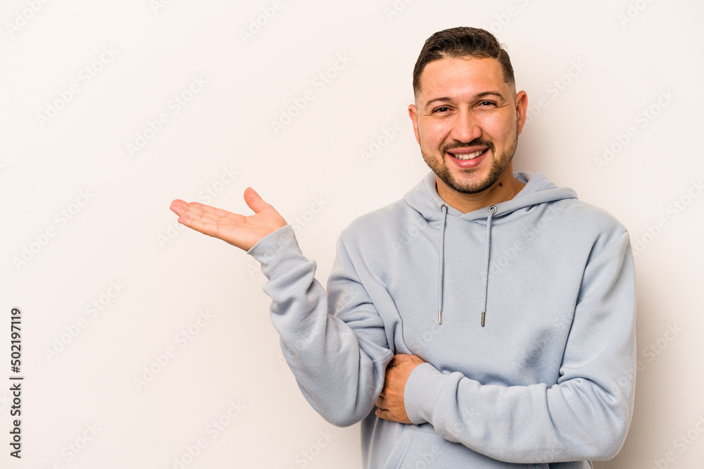 Young hispanic man isolated on white background showing a copy space on a palm and holding another hand on waist. Young hispanic man isolated on white background showing a copy space on a palm and holding another hand on waist.