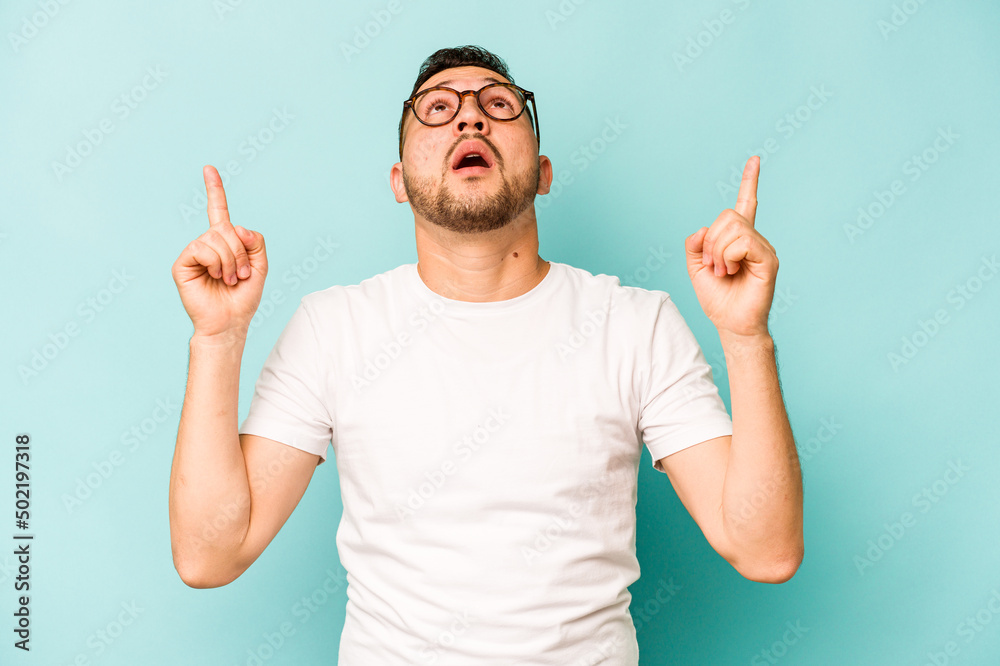 Young hispanic man isolated on blue background pointing upside with opened mouth.