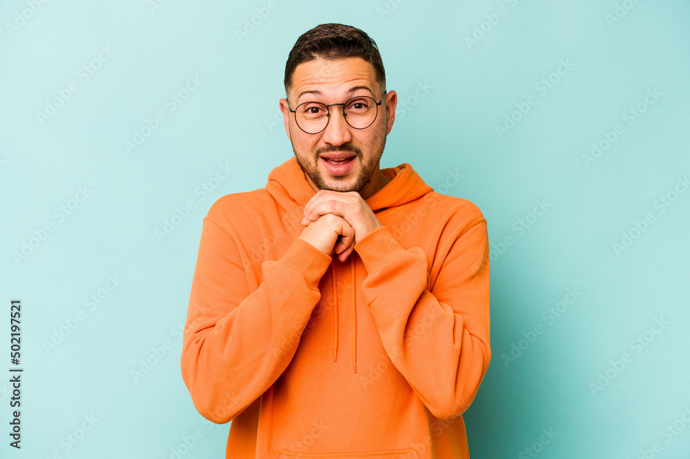 Fototapeta premium Young hispanic man isolated on blue background praying for luck, amazed and opening mouth looking to front.