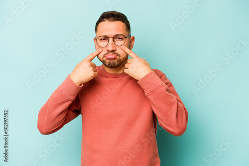 Young hispanic man isolated on blue background crying, unhappy with something, agony and confusion concept.