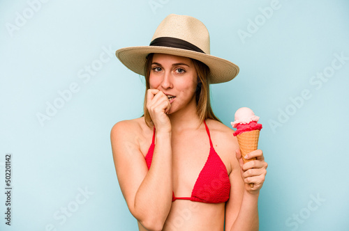 Young caucasian woman wearing a bikini and holding an ice cream isolated on blue background biting fingernails, nervous and very anxious.