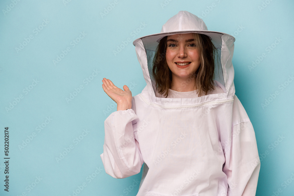 Young caucasian beekeeper woman isolated on blue background showing a copy space on a palm and holding another hand on waist. Young caucasian beekeeper woman isolated on blue background showing a copy space on a palm and holding another hand on waist.