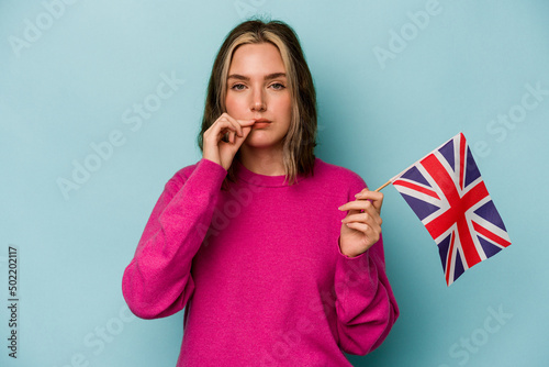 Young caucasian woman holding a English flag isolated on blue background with fingers on lips keeping a secret.