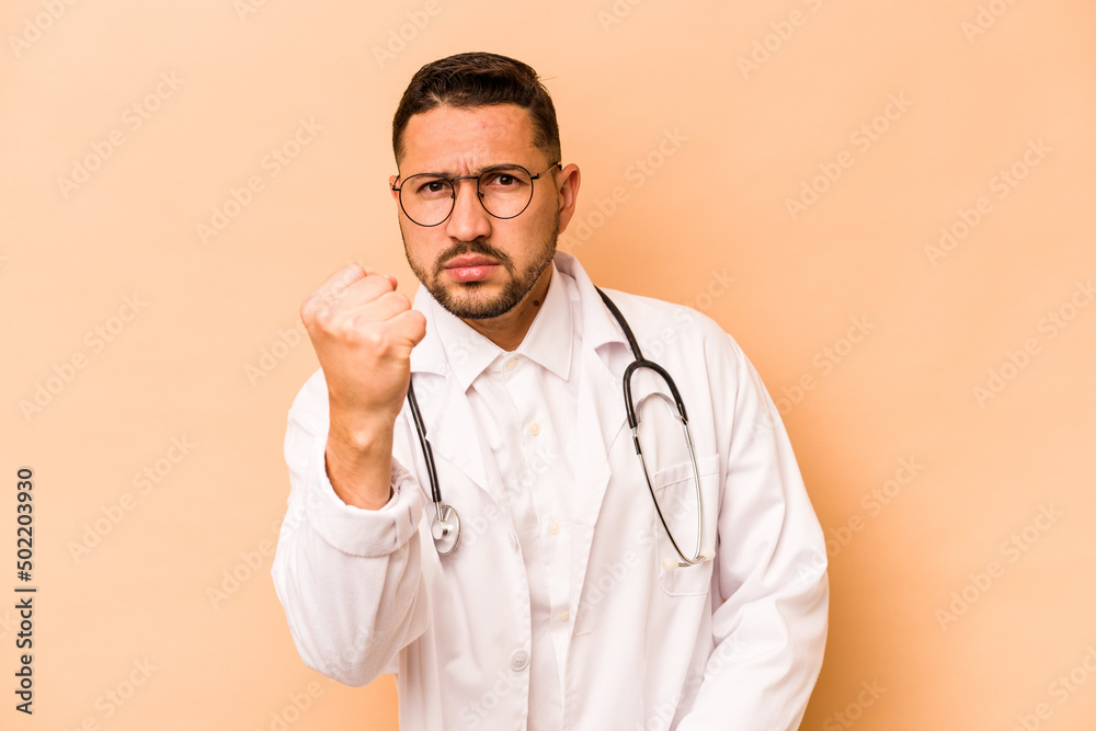 Hispanic doctor man isolated on beige background showing fist to camera, aggressive facial expression. Hispanic doctor man isolated on beige background showing fist to camera, aggressive facial expression.