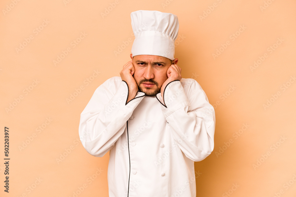Hispanic chef man isolated on beige background covering ears with hands. Hispanic chef man isolated on beige background covering ears with hands.