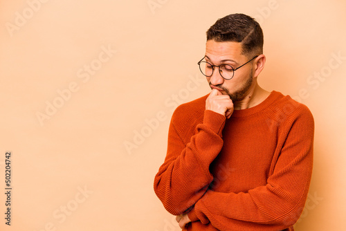 Young hispanic man isolated on beige background looking sideways with doubtful and skeptical expression.