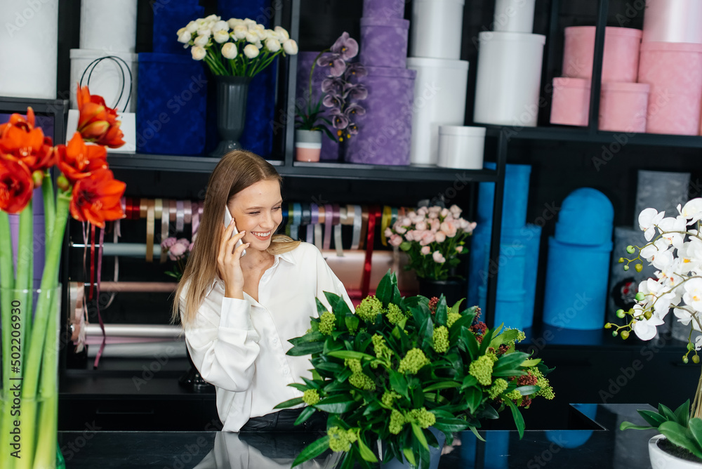 Fototapeta premium A young woman florist accepts an order by phone for the production of a beautiful festive bouquet. Floristry and making of bukets in a flower shop. Small business.