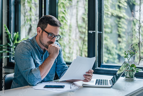 Anxious businessman analyzing documents while working on laptop at table