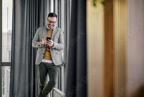 Smiling businessman networking on smart phone while standing by window at office