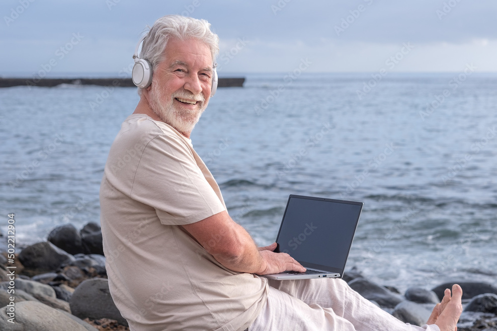 Smiling bearded man sitting barefoot on pebble beach wearing headphones while using laptop. Elderly happy man looking at camera enjoying remote work