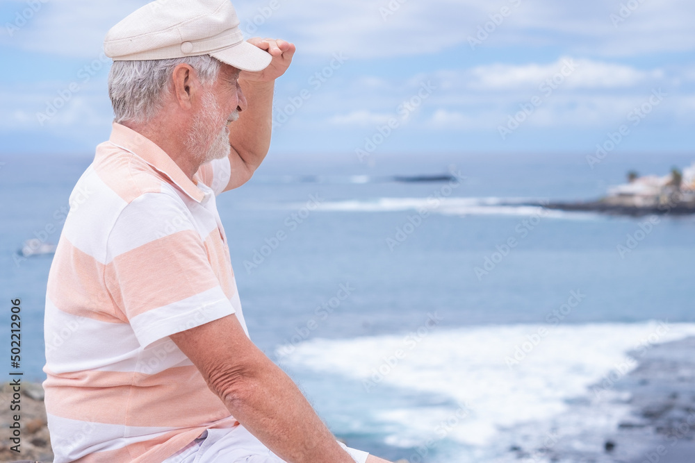 Attractive senior man with beard and hat sitting outdoors at sea looking at horizon enjoying freedom and vacation. Active caucasian elderly man relaxing on a sunny day close to the beach
