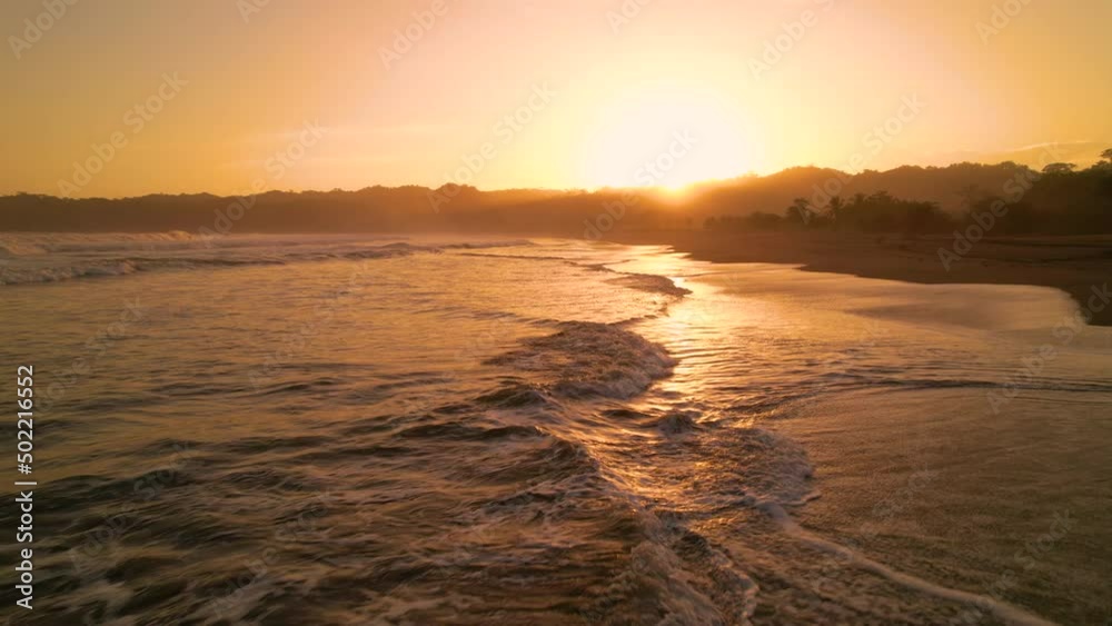 AERIAL: Ocean waves spilling on a tropical sandy beach in golden light. Dazzling sun reflection on an empty tropical shoreline in Panama. Peaceful and serene summer moment at Playa Venao.