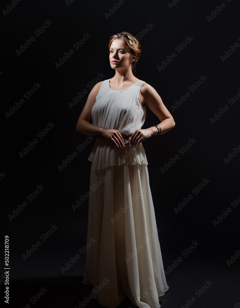 Fototapeta premium An ancient heroine, a young woman in the image of an ancient Greek goddess or muse. A noble heroine in a white tunic and a laurel wreath, a full-length photo on a black background