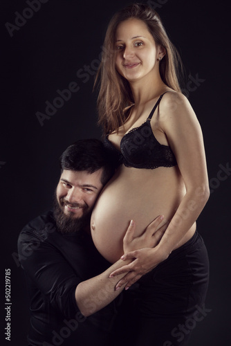 a married couple, a man with a beard and a pregnant woman with a big belly, a man listens to her belly, smiling