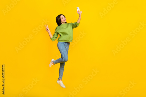Full length photo of young girl jump shooting selfie mobile show peace v-symbol isolated over yellow color background